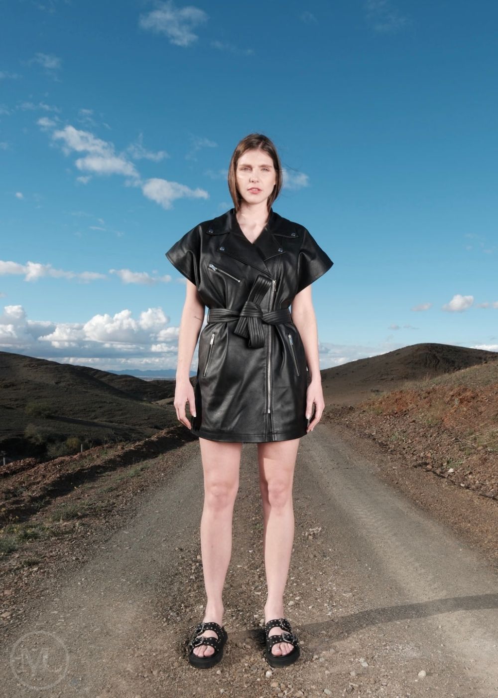 Woman in a black leather dress in biker style standing on a desert road with a blue sky.