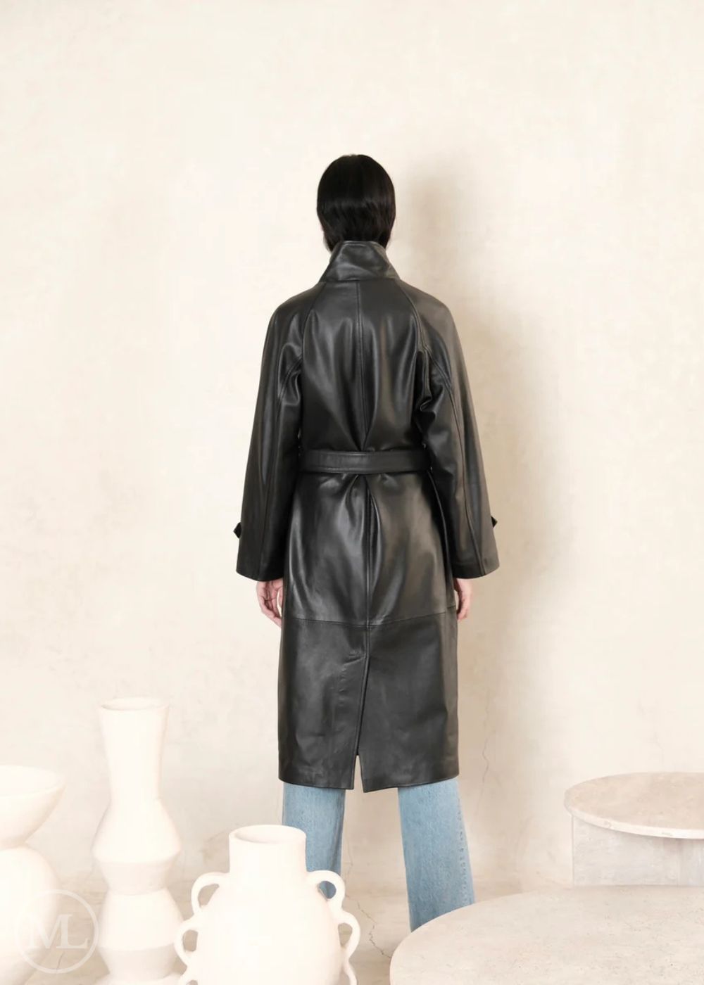 women standing backward wearing black leather trench coat with belted waist and a beige background and back view of coat